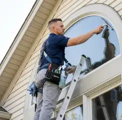 Professional window cleaner on a ladder using a squeegee to clean an exterior arched window, Riverside, CA.